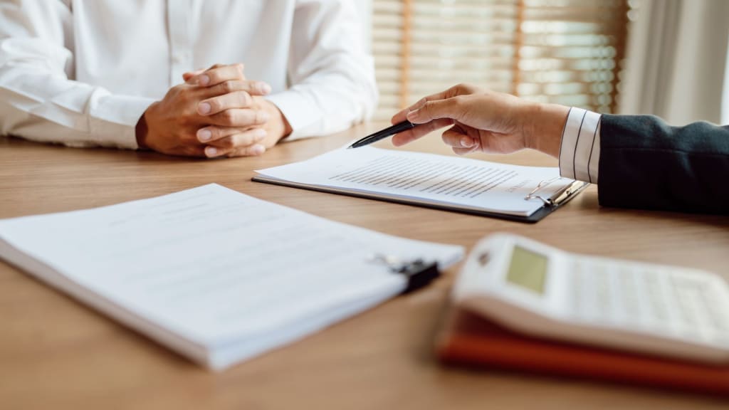 Two people sit at a desk with documents, a clipboard, and a calculator; one person points at a form with a pen while the other has their hands clasped.